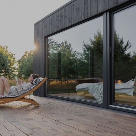 Eine Person, die sich während des Sonnenuntergangs auf einem Liegestuhl am großen Fenster entspannt, mit Bäumen im Hintergrund.